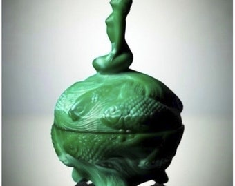 Puede incluir: Una caja de vidrio verde para guardar pequeños objetos con una figura de sirena en la parte superior. La caja está decorada con un patrón de peces y algas marinas en espiral.
