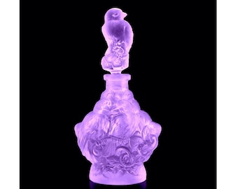 May include: A lavender-coloured glass perfume bottle with a bird-shaped stopper. The bottle has a detailed, ornate design with a sculpted base. The background is black.