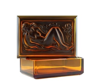 May include: A rectangular amber glass box with a raised design of a nude woman reclining on a bed of leaves. The box is open and the lid is resting on top of the box.