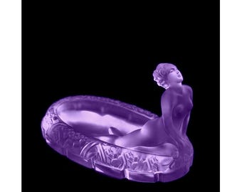 May include: A purple glass ashtray featuring a sculpted female figure reclining on the rim. The figure is nude and has a stylised hairdo. The ashtray is decorated with floral motifs.