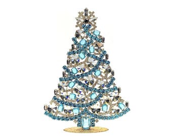 May include: A decorative tabletop Christmas tree ornament constructed from a metal frame encrusted with numerous light blue, dark blue, and clear faceted crystals. The tree features a star-shaped topper and stands on a circular gold base.