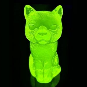 May include: A luminous green cat ornament with a textured finish. The cat is depicted in a seated position, with its ears erect and eyes closed. The background is a deep black, accentuating the vibrant green hue of the cat.