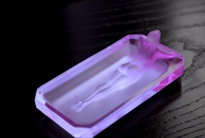 K&ouml;nnte beinhalten: Ein Aschenbecher aus violettem Glas mit einem erhabenen Design einer liegenden Person.