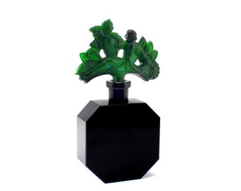 Puede incluir: Una botella de vidrio negro con un tapón de vidrio verde que presenta una figura estilizada. El tapón tiene la forma de una mujer sentada en una rama con hojas.