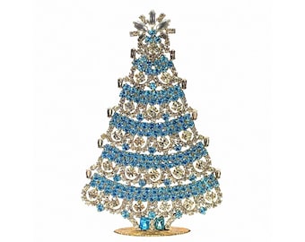 May include: A decorative tabletop Christmas tree ornament constructed from a metal frame encrusted with alternating horizontal bands of clear and light blue rhinestones, featuring a star-shaped topper and a circular base stand.