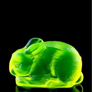 May include: A translucent, vibrant lime green glass figurine shaped like a resting rabbit. The sculpture features smooth, rounded contours and a polished surface that catches the light, highlighting the rabbit's ears and curled body posture.