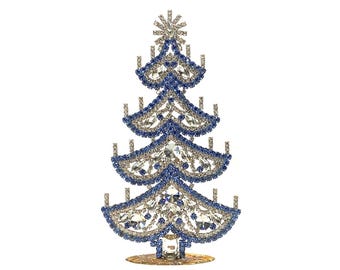 Op de afbeelding: Een decoratief kerstboomfiguur. De boom is gemaakt van zilverkleurig metaal en is versierd met blauwe en heldere kristallen. Hij heeft een ster op de top en een goudkleurige basis. De boom is ongeveer 20 cm hoog.