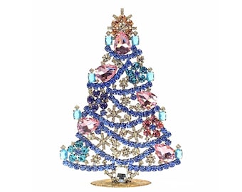 Pode incluir: Uma figura decorativa de árvore de Natal. A árvore é adornada com pedras preciosas azuis, rosas e transparentes, dispostas em um design festivo. A base é dourada, em um fundo branco.