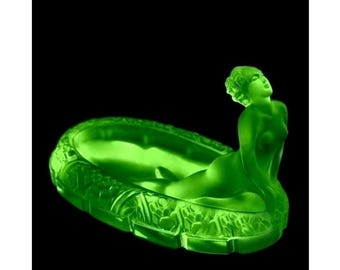 May include: A vibrant green glass dish featuring a sculpted figure of a reclining person at one end. The oval-shaped vessel has a textured, relief-patterned exterior rim and a smooth, translucent finish that glows with a bright, vivid green hue.