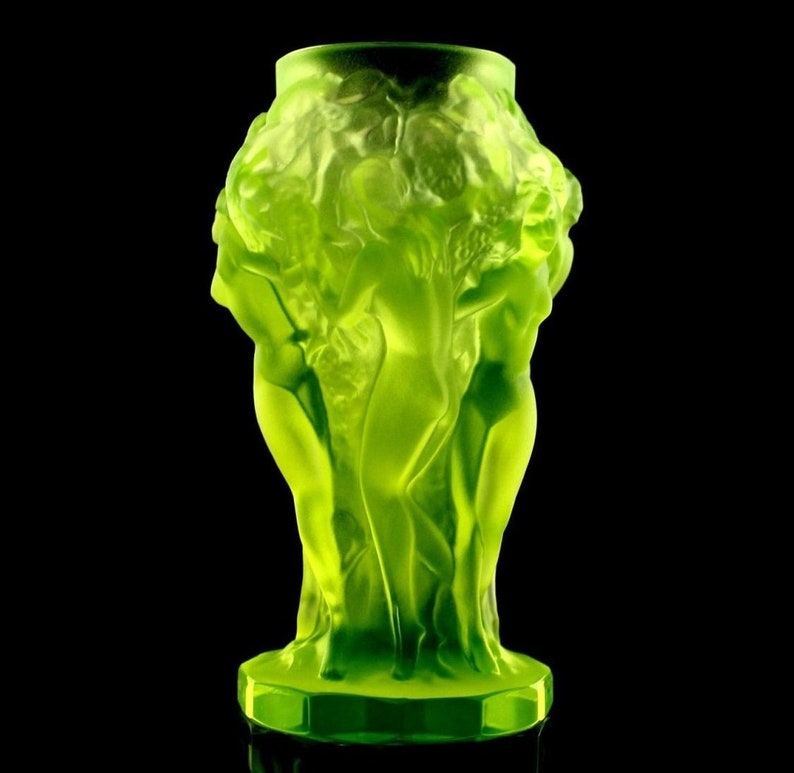 May include: A green glass vase with a design of three women standing together. The vase is made of frosted glass and has a textured surface.