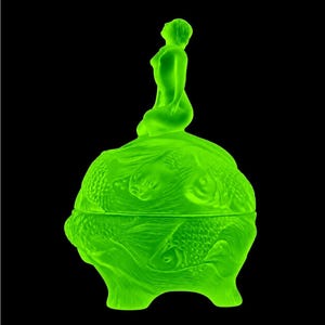 Op de afbeelding: Een levendige neongroene glazen pot met een gebeeldhouwd deksel bekroond door een zittende figuur die omhoog kijkt. Het lichaam van de pot is versierd met reliëfs van meerdere vissen die in een cirkelvormig patroon zwemmen, wat een textuur creëert.