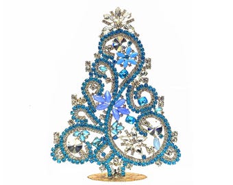 May include: A decorative Christmas tree figurine. The tree is adorned with blue and silver rhinestones and gemstones. The tree is set on a gold-colored base and has a star-shaped topper. The tree is a festive holiday decoration.