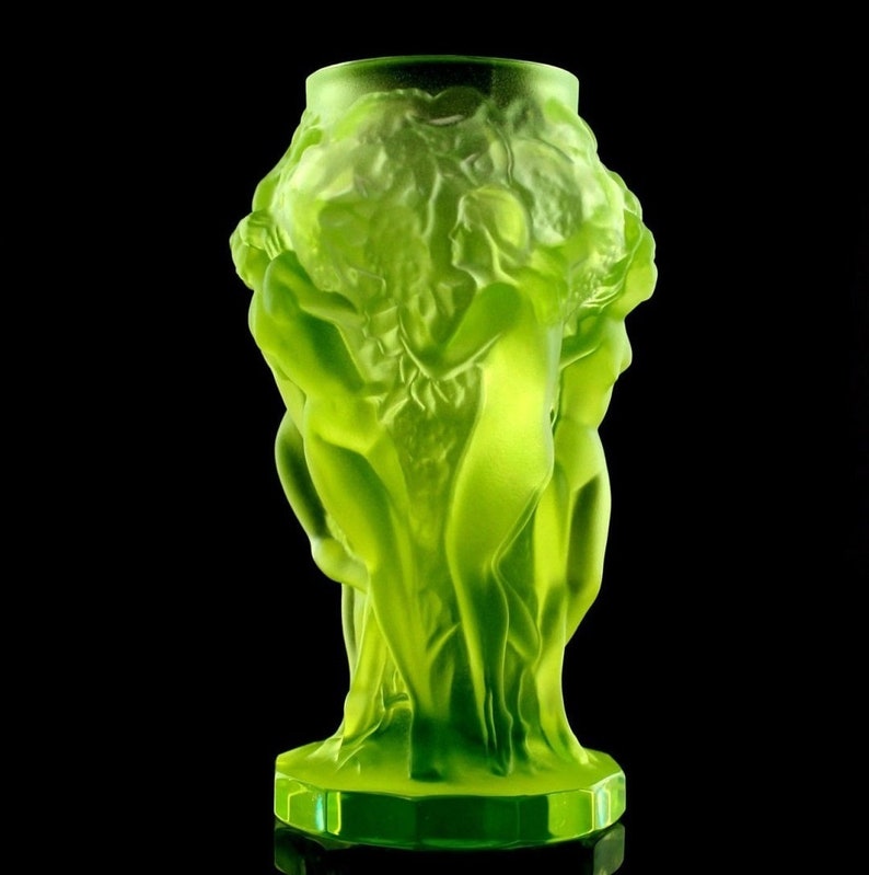 May include: A green glass vase with a design of three figures intertwined. The vase has a textured surface and a hexagonal base.