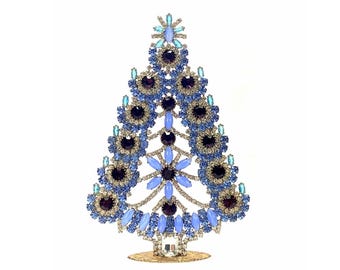 May include: A decorative Christmas tree ornament crafted from numerous blue, light blue, and dark purple rhinestones set in a silver-toned metal frame. The tree features a star-shaped top, layered circular patterns, and a circular base stand.