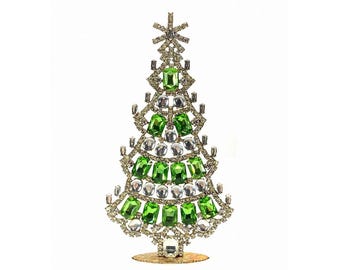 May include: A decorative Christmas tree figurine. The tree is made of gold-toned metal and adorned with green and clear gemstones. The tree has a star on top and sits on a round base. The gemstones are arranged to resemble a Christmas tree.