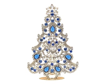 May include: A decorative Christmas tree ornament crafted from silver-toned metal filigree, intricately set with numerous clear white rhinestones and vibrant blue teardrop and round-cut crystals, standing on a small circular base.