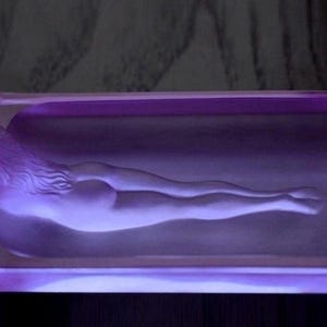 K&ouml;nnte beinhalten: Ein Aschenbecher aus violettem Glas mit einem Relief einer liegenden nackten Figur.