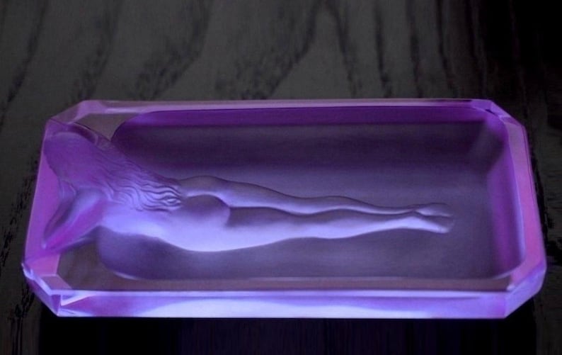 K&ouml;nnte beinhalten: Ein Aschenbecher aus violettem Glas mit einem Relief einer liegenden nackten Figur.
