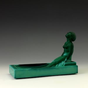 Puede incluir: Un cenicero de vidrio verde con una figura esculpida de una mujer recostada en el borde. La mujer está desnuda y tiene la cabeza echada hacia atrás, mirando hacia arriba.