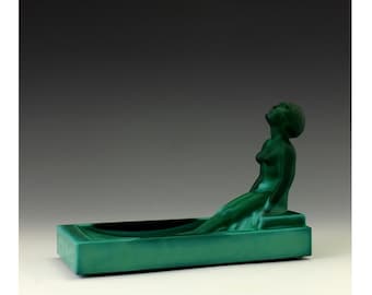 Puede incluir: Un cenicero de vidrio verde con una figura esculpida de una mujer recostada en el borde. La mujer está desnuda y tiene la cabeza echada hacia atrás, mirando hacia arriba.