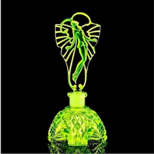 May include: A green glass bottle with a decorative stopper featuring a stylised female figure. The bottle has a faceted design and a round base.