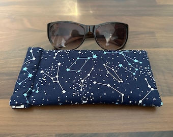 May include: A navy blue glasses case with a constellation pattern. The case has white stars and constellations on a dark blue background. The case is closed and a pair of brown sunglasses are sitting on top of it.