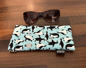 May include: Black sunglasses rest above a light blue glasses case. The case is patterned with black and white penguins. The case has a small black tag with the word "moo" on it.