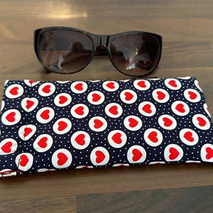 May include: A navy blue glasses case with a white polka dot pattern and red hearts. The case is closed and lying on a wooden surface.
