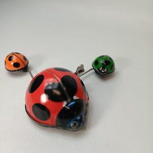 May include: A red ladybug-shaped pin with black spots and two smaller ladybugs on either side. One is orange with black spots, and the other is green with black spots. The pin is on a white background.
