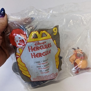 May include: A sealed plastic bag containing two Disney's Hercules Happy Meal toys. One is a figurine of Phil, and the other is a figurine of Nessus. The packaging features the McDonald's logo and the text "Disney's Hercules".