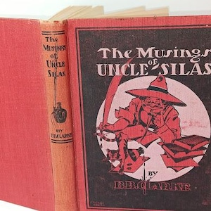 May include: An open antique book titled "The Musings of Uncle Silas" by B.B. Clarke. The cover is red with a black and red illustration of a man in a hat. The spine is orange with the title and author's name.