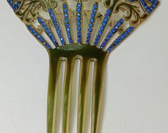 May include: An Art Nouveau style hair comb with a fan-shaped top. The comb features a gold-toned filigree design adorned with blue rhinestones. The teeth of the comb are a light green color.