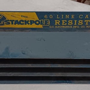 Op de afbeelding: Vintage blauwe metalen Stackpole Resistors display. Het bovenste gedeelte heeft een geel en blauw label met de tekst "G&L STACKPOLE" en "60 LINE CARBON RESISTORS". De display heeft vier horizontale sleuven voor het opbergen van weerstanden. De tekst "G.C. ELECTRONICS MFG. CO. ROCKFORD, ILL. U.S.A." is ook zichtbaar.