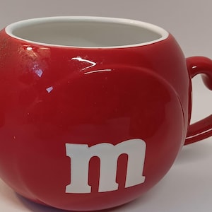 May include: Red ceramic mug with a white "m" on the front and a heart-shaped handle.