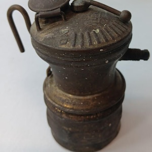 Könnte beinhalten: Eine antike Öllampe aus Messing mit rundem Sockel und gewölbtem Deckel. Die Lampe hat einen Griff, einen kleinen Deckel und eine seitliche Düse. Das Metall hat ein verwittertes Aussehen, das auf Alter und Gebrauch hindeutet. Die Lampe ist ein Vintage-Artikel.