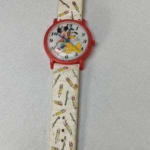 May include: A vintage wristwatch with a red case and a white face featuring Mickey Mouse and Pluto. The watch band is cream-colored and decorated with yellow pencils. The watch is a fun, nostalgic accessory.
