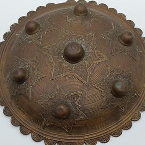 Peut inclure: Un plateau circulaire en métal de couleur bronze avec un bord festonné et un dôme central surélevé. La surface est ornée de six dômes plus petits, chacun entouré d'un motif gravé en forme d'étoile à huit branches.