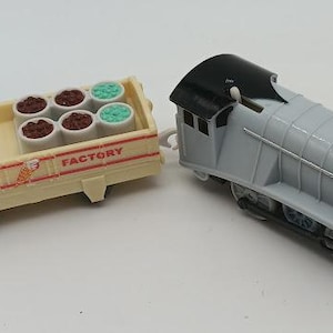 Könnte beinhalten: Eine graue und schwarze Spielzeug-Dampflokomotive, die an einen cremefarbenen Güterwagen angehängt ist. Der Wagen trägt die rote Aufschrift ICE CREAM FACTORY und enthält sechs kleine runde Behälter, vier braune und zwei hellgrüne, die Eis darstellen.