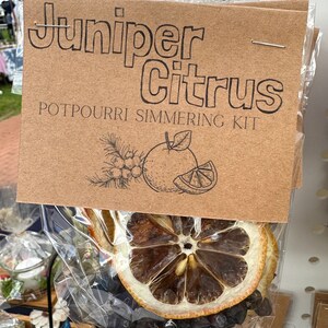Puede incluir: Una bolsa de papel marrón con el texto "Juniper Citrus Potpourri Simmering Kit" y un dibujo de una fruta cítrica. La bolsa contiene rodajas de cítricos secos y otros ingredientes.