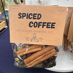 Peut inclure: Un sac en papier brun avec une étiquette indiquant "Spiced Coffee Potpourri Simmering Kit". Le sac contient des bâtonnets de cannelle, des grains de café et d'autres épices.