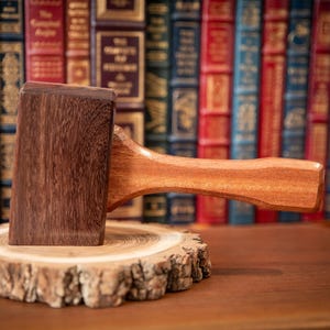 May include: A wooden gavel with a dark brown head and a lighter brown handle rests on a small, round wooden slice. The background features a collection of antique books with various colored spines, suggesting a legal or academic theme.