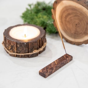 May include: A lit candle in a wooden holder with a natural bark finish, next to a wooden incense holder with a lit incense stick. The incense holder is a rectangular piece of wood with a natural wood grain.