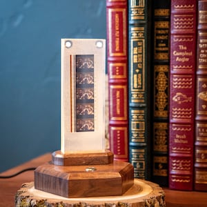 May include: A unique film strip lamp with a wooden base and a clear acrylic frame. The lamp features a vintage film strip and is set on a wooden base, resting on a natural wood slice. Books with gold lettering are in the background.