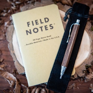 May include: A brown wooden pen with a silver tip and a black pen pouch. The pen is next to a yellow notebook with the text "FIELD NOTES" and "48-Page Memo Book - Durable Materials / Made in the U.S.A."