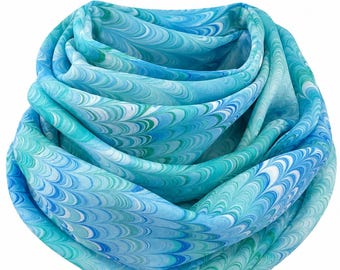 May include: A lightweight scarf featuring a marbled pattern in shades of turquoise, sky blue, royal blue, and white. The fabric is arranged in soft, layered folds, creating a continuous wave-like design across the entire surface of the material.
