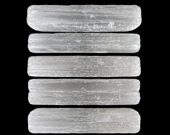 May include: Five translucent, light gray selenite crystal wands are arranged horizontally against a black background. The wands have a striated texture and rounded ends, showcasing their natural crystalline structure. These wands are used for spiritual practices.