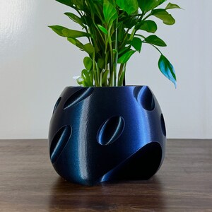 May include: A dark blue, spherical planter with an abstract design, filled with vibrant green leafy plants. The planter has a textured surface with oval-shaped cutouts. The plant pot is sitting on a wooden surface.