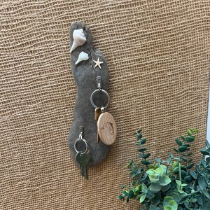 May include: A gray stone key holder shaped like a foot, adorned with seashells and a starfish. It features metal hooks holding keys and a wooden key tag. The key holder is mounted on a textured, tan background.