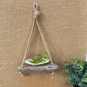May include: A rustic wooden shelf with a natural rope hanger, holding a small succulent plant in a white dish. The shelf is hanging against a burlap background.