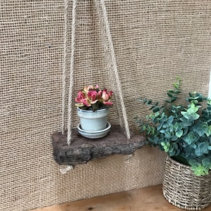 May include: A small wooden shelf with a rope hanging system, holding a potted succulent plant with pink flowers. The shelf is made from a natural piece of wood bark and is hanging against a burlap background.